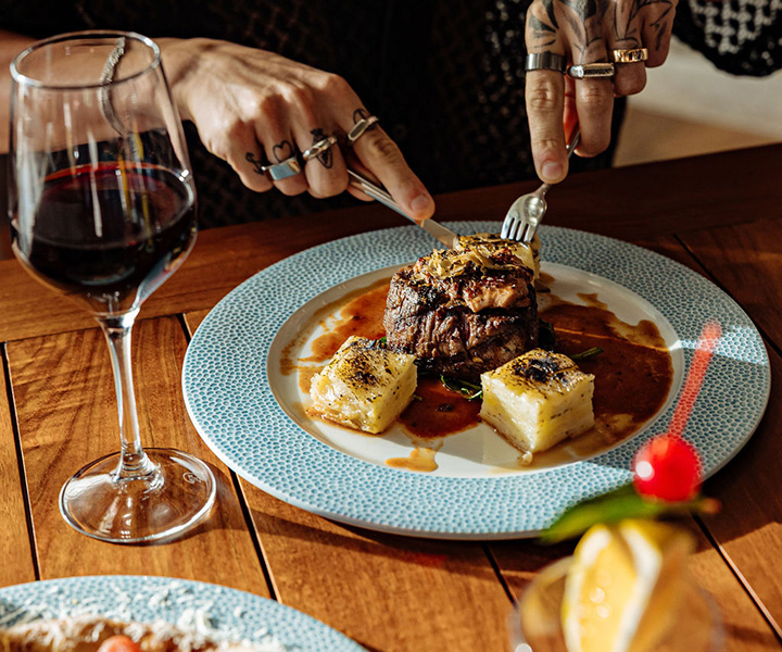 A man cutting into a hearty meat dish with a glass of red wine