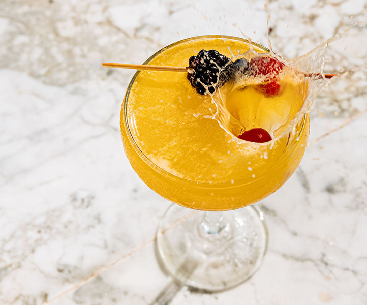 Yellow cocktail with a blackberry garnish and an ice cube splashing in it