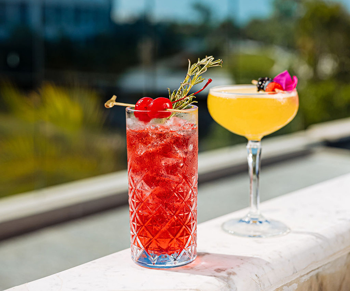 two cocktails, one red in a collins glass and the other yellow in a coupe, sitting outside in the sunshine