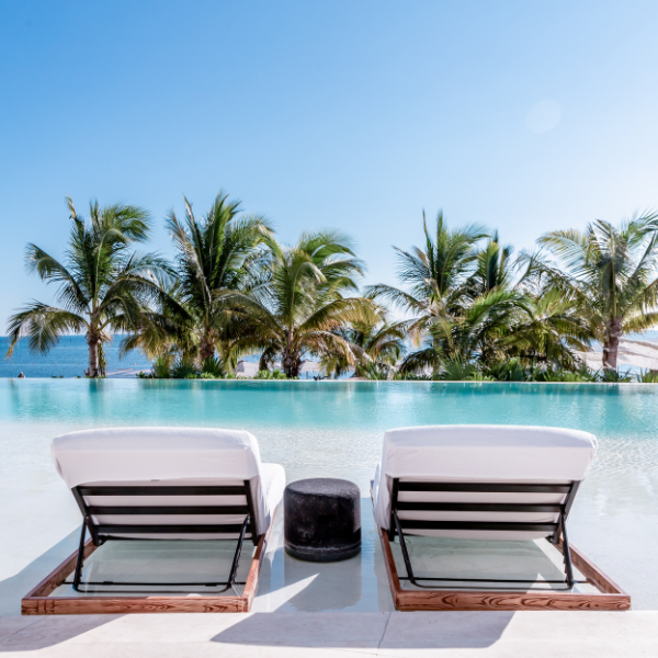 two luxurious lounge chairs sitting next to a beach entrance style infinity pool facing palm trees